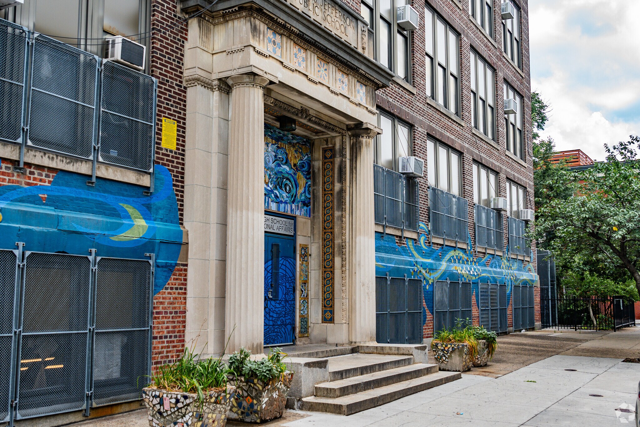 Main entrance of Bodine High School featuring stone columns, blue doors, colorful mosaic artwork, and blue murals along the brick exterior.