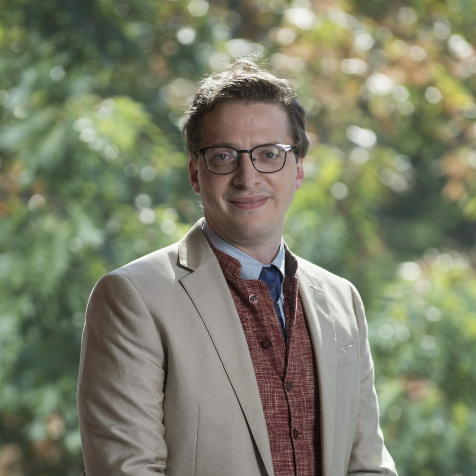 Principal Dr. Brown stands outdoors wearing glasses, a tan blazer, and a red patterned vest, with trees in the background.